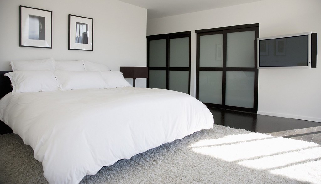 A white bed in a plain room, with low sun across a pale rug